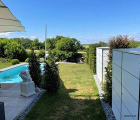 Ein moderner Garten mit kleinem Pool, Wiese, Pflanzen, Staue und Sichtschutz unter blauem Himmel.