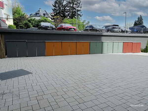 Kleine Schränke an einer Wand, bunte Türen, Pflastersteine, Autos auf erhöhtem Parkplatz im Hintergrund.