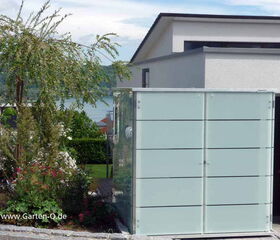 . The image shows a modern outdoor setting. There is a structure that appears to be a garden storage unit with frosted glass panels. To the left, there is a small garden with various plants and a tree. In the background, there is a building with a flat roof, and part of a white vehicle is visible. The website URL "www.Garten-Q.de" is on the image.