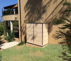 . The image shows a modern outdoor area with a tan house wall. There's a rectangular box or structure against the wall, possibly a storage unit. A balcony with plants is visible above, supported by a white column. The foreground has green grass and some bushes, while trees and plants are in the background, including a tree with fern-like leaves on the right. The area seems open and sunny.
