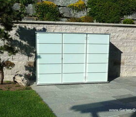 . The image shows a minimalist outdoor storage unit with a frosted glass or light-colored panel design, set against a stone wall. There's a tree and some greenery on one side, and the unit is placed on a stone or tiled patio area.