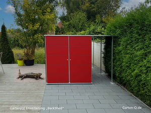 Ein rotes Gartenhaus mit einer Eidechse davor auf einer Terrasse, umgeben von Pflanzen.
