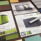 The image shows a variety of printed marketing materials and color samples arranged on a flat surface. There are brochures or flyers featuring images and text, possibly related to garden products or architectural designs, as suggested by the term "Garten[Q]" on one of the brochures. A small ruler is placed on top of one of the brochures. The materials display images of structures with various color options.