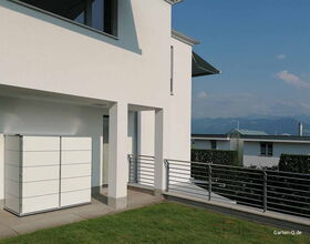 Ein modernes Haus mit Terrasse, Geländer und einem weißen Schrank im Freien. Hintergrund zeigt Berge.