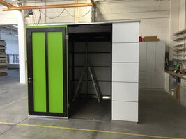 The image shows a compact room or booth with a green door standing open in what appears to be an industrial or workshop setting. Inside, there is a ladder leaning against the back wall. The surrounding area includes cabinets, shelves, and various equipment, indicating it might be part of a workspace or storage area.