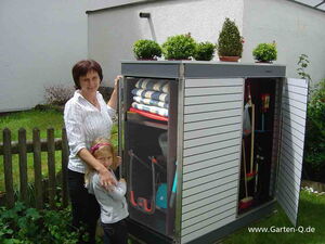 . The image shows a woman and a child standing next to an outdoor storage cabinet. The cabinet is open, displaying various garden tools and supplies inside, such as brooms, rakes, and folded towels. On top of the cabinet, there are several potted plants. The setting appears to be a garden or backyard.