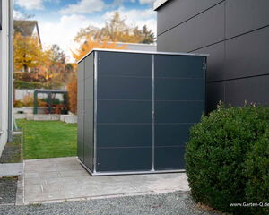 . The image shows a modern, sleek outdoor storage shed placed on a paved area next to a building. The shed has a rectangular shape with a dark finish and metallic edges. To the right, there's a bush, and behind the shed is a lawn leading to a landscaped area with trees in the background. The image also contains the URL "www.Garten-Q.de" at the bottom right corner.