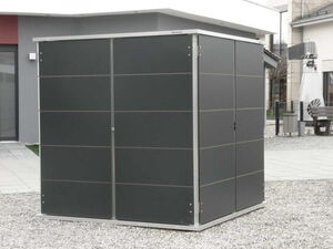 . The image shows a dark-colored metal shed with a modern design. It's located outdoors on a gravel surface, and there are buildings visible in the background. The shed has a sleek appearance with vertical lines and a flat roof.