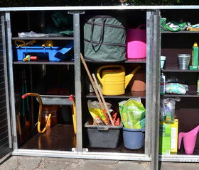 . The image shows a garden storage shed or cabinet with open doors. Inside, there are various gardening tools and supplies, including:

1. Watering cans
2. A wheelbarrow
3. Hand tools
4. Buckets
5. Plant pots
6. Garden clogs
7. Shelves with gardening items like fertilizer, seeds, and possibly pest control products
8. A storage bag and trays

This setup is organized for easy access to gardening essentials.
