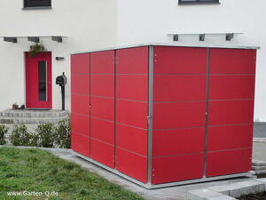 . The image shows a modern outdoor storage unit or shed with a sleek design featuring red panels and a metal frame. It is placed in a yard area, next to a house with a matching red door. The scene includes a small lawn, some paved walking areas, and a mailbox near the entrance of the house.