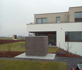 . This image shows a modern two-story house with a simple geometric design. The exterior is painted in neutral colors, and large windows are present. The yard has a small patch of grass and a paved area with what appears to be an outdoor structure or enclosure. There's also gardening equipment visible on the lawn. The sky is overcast, suggesting an overcast day.
