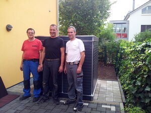 . The image shows three men standing side by side on a paved walkway next to a large, rectangular, dark-colored unit. Behind them is a light yellow building wall, and a green area with bushes and trees. The men are dressed in casual workwear and appear to be smiling.