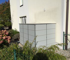 . The image shows a rectangular metal structure, possibly a storage unit or enclosure, situated next to a building. It's surrounded by some landscaping, including flowering shrubs and other greenery. The area is enclosed by a metal fence, and the building exterior is light-colored. The scene is well-lit, indicating it's daytime.