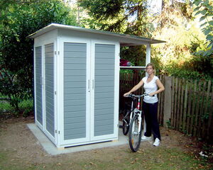 The image shows a small outdoor storage shed with double doors. A person is standing next to it with a bicycle. The shed is situated on a concrete slab, and there are trees and a wooden fence in the background.