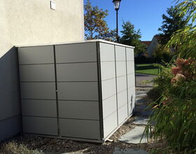 . The image shows a modern, metal storage shed located next to a building. It is rectangular in shape and appears to be on a bed of small gravel and flat stepping stones. There are some plants and a lamppost in the background, with a clear blue sky above.