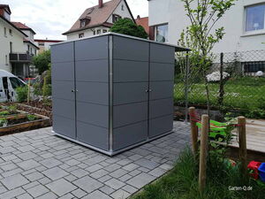 . Ein moderner, grauer Gartenschuppen auf einem gepflasterten Bereich, umgeben von Garten und Häusern.
