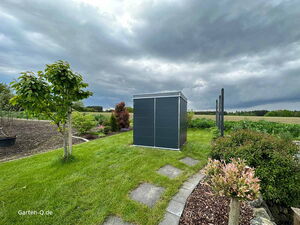 . Ein Garten mit Rasen, Bäumen, Pflanzen und einem dunklen Gartenschrank unter bewölktem Himmel.