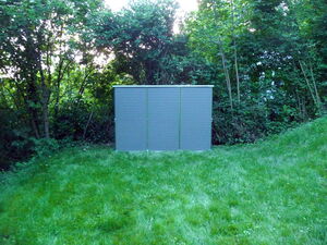 . The image appears to show a metal cabinet or shed placed on a grassy area with trees and foliage in the background.
