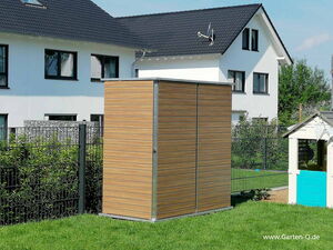 . The image depicts a garden shed with a modern, sleek design featuring wood paneling and metal framing. It's positioned on a grassy area in front of a fence. In the background, there's a two-story house with a dark roof and white walls. There's also a small playhouse with a blue door on the right side.