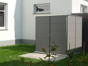. Ein grau-grüner Gartenaufbewahrungsschuppen steht auf einem Rasen neben einem weißen Gebäude.