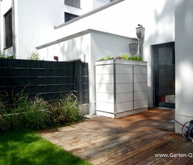 . In the image, there is a patio area with wooden decking, adjacent to a grassy lawn. There's a storage cabinet with plants on top against the wall of a modern white building. A black fence and some tall grass plants are on the left. Additionally, a garden hose is coiled on the right side of the image. The setting appears to be a well-maintained outdoor space.