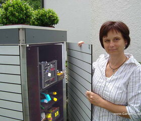 . The image shows a person standing next to an outdoor storage cabinet. The cabinet is open, revealing shelves that contain various items such as a crate, some tools, and a few other objects. On top of the cabinet, there are two potted plants. The setting appears to be outdoors, possibly in a garden or backyard area.