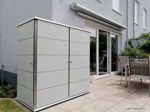 . The image shows a modern backyard setup. There's a large, sleek storage unit or shed on a patio next to a house with sliding glass doors. A table with chairs is set up nearby for outdoor dining. The area is neat and well-maintained, with some greenery and plants visible.