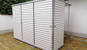 . Ein moderner, weißer Gartenschrank mit Lamellen, steht auf einem gepflasterten Bereich vor grüner Hecke.