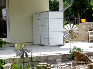 . The image shows a patio or garden area. There is a modern storage cabinet against the wall, with a sleek, white and gray design. In the foreground, several garden ornaments resembling silver wind spinners or decorative pinwheels are visible. There's also outdoor furniture in the background, a drainage pipe on the house, and stone landscaping elements like small rocks and a retaining wall.