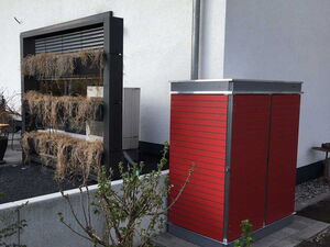 . The image shows an outdoor setting with a red vertical structure, likely a shed or storage unit. Next to it is a vertical garden with several tiers, each containing dry or withered plants. The garden is situated near a wall, and there's a pipe running alongside the building. There are also some small plants or bushes in the foreground.