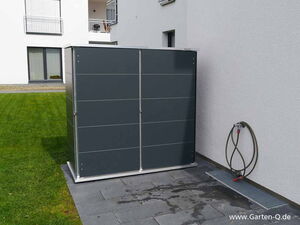 . Eine graue Gartengerätebox steht auf einer Terrasse neben einer Hauswand, mit einem Wasserschlauch an der Wand.
