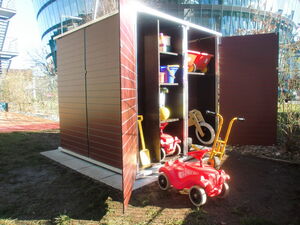 . The image shows an outdoor storage shed with its doors open, revealing a variety of items inside. There are shelves containing bright-colored toys including a ride-on car and buckets. A toy wheelbarrow and a wooden balance bike are also visible. Additionally, you can see gardening tools like a yellow shovel. The storage unit is situated outside, with a modern building and trees in the background.