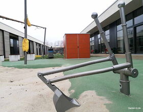Ein Sandkastenbagger auf einem Spielplatz vor einem modernen Gebäude mit gelben Stofftaschen an Stangen.