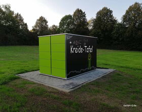 . Ein grüner Gartenschrank mit Kreidetafel, steht auf einem gepflasterten Platz in einer Wiese.
