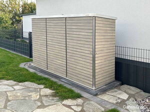 . Ein Gartenschrank aus Holzpaneelen neben einem Zaun und einer Steinfliesenfläche.