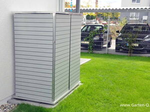 . Ein moderner Gartenschrank aus Metall steht neben einem Haus auf einer grünen Rasenfläche. Im Hintergrund parken Autos.