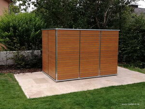 . This image shows a wooden garden shed with a modern design. The shed is placed on a concrete slab and is surrounded by a garden area with grass and trees in the background. The wood panels have horizontal lines and the structure is bordered with metal frames.