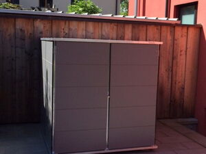 . The image shows a gray storage shed placed on a paved area outside. It is positioned against a wooden fence, with buildings visible in the background.