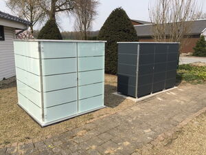 . The image shows two rectangular storage units or cabinets outdoors. They are sleek and modern in design, with smooth surfaces and horizontal lines. One is light-colored, while the other is dark. They are placed on a paved and gravel area with trees and shrubs in the background.