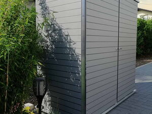 . Ein graues Gartenhaus aus Metall steht neben Bambus und einem Steinweg, unter einem bewölkten Himmel.