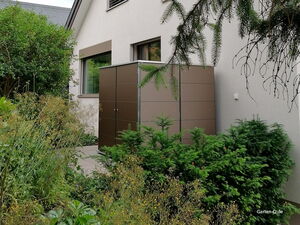 . Ein brauner Gartenschrank steht neben einem Haus in einem üppig begrünten Garten.