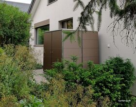 . Ein moderner Gartenschrank neben einem Haus, umgeben von Pflanzen und Büschen.