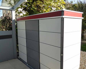 . The image shows a modern storage unit, likely for garden tools or bicycles. It has a sleek design with horizontal panels in shades of gray and white. The unit is placed under a transparent canopy, and there are plants and a house visible in the background. The design looks contemporary and functional.