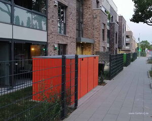 The image shows a modern residential street with apartment buildings. The buildings feature brick exteriors and large windows. In the foreground, there are metal fences and an orange structure, possibly for storage or utilities. The sidewalk runs parallel to the buildings, lined with some greenery and trees.