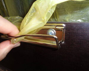 . The image shows a person holding a yellow plastic bag that appears to be attached to a holder or frame on a dark-colored surface. The holder seems to be metal and is screwed into the surface. The setup looks like it's for securely holding a trash bag in place.