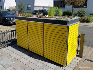 . Gelber Gartenschrank mit Gründach und Pflanzen, steht auf einer Pflasterfläche in einer Wohngegend.