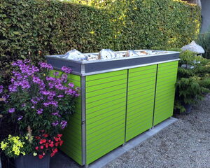 . The image shows a green storage unit or enclosure with a modern design, featuring horizontal slats. It is placed outdoors on a gravel surface. To the left of the unit, there's a pot with various colorful flowers, including purple, red, and yellow blossoms. The background includes a well-trimmed hedge and more plants.