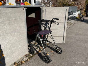. Ein Rollator steht vor einem offenen Holzschrank auf einem Weg, daneben sind zwei Fahrräder.