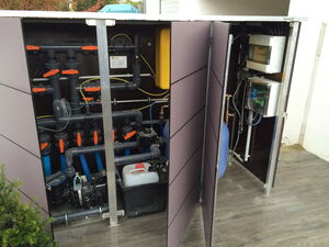 . The image shows an equipment enclosure with various components likely related to a pool or water system. It contains:

1. Plumbing pipes and fittings with orange-handled valves.
2. A pump or motor unit.
3. Electrical components or control boxes mounted on the side.
4. A canister or container, possibly for chemicals.
5. Wiring connecting different parts.

The setup looks organized for managing a water system.