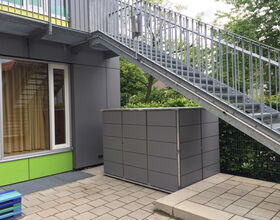 . The image shows:

1. A metal staircase positioned above a boxed structure.
2. A large window with curtains beside the structure.
3. Gray panels on the walls and box, possibly storage or utility covering.
4. Paved stone flooring.
5. Some greenery and trees in the background, creating a natural backdrop.